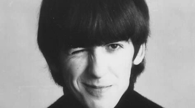 A black and white close-up photo of George Harrison in the early days of the Beatles. He is smiling a closed-mouth smile and winking. He wears a black high-collared sweater and has a "mod" haircut with bangs.