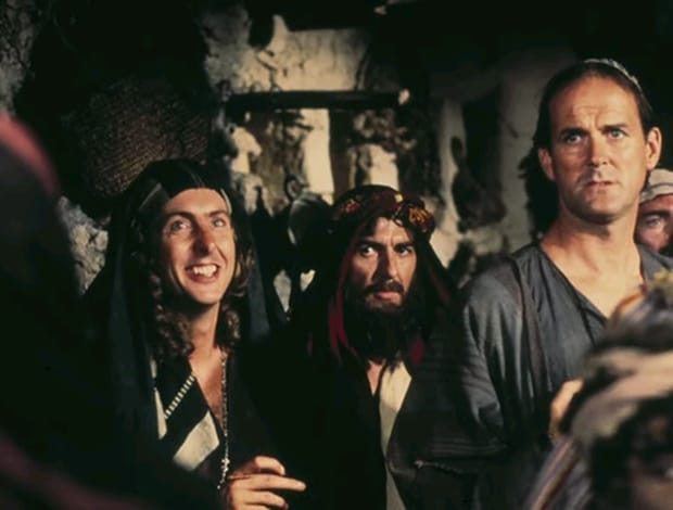 A scene from Monty Python's Life of Brian with a smiling Eric Idle in a striped head-covering shawl; a serious-looking George Harrison, also in a shawl, with long hair, moustache, and beard; and John Clease in a simple grey shirt and a yarmulke.