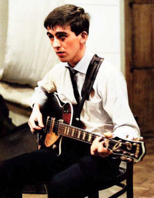A young George Harrison with short hair and a big black eye sits on a black chair and looks off to the left. He holds an electric guitar, and wears a white collared shirt, black skinny tie, and black skinny trousers. 