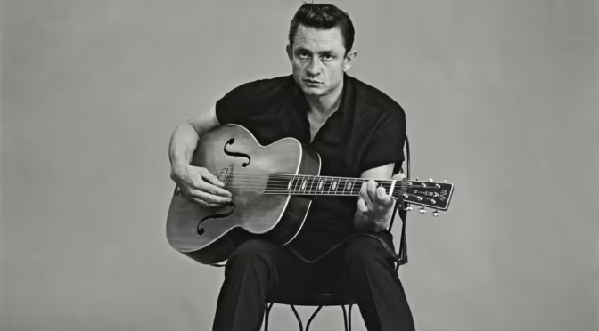 A black and white photo of Johnny Cash dressed in black sitting on a stool playing an acoustic guitar in front of a white background.