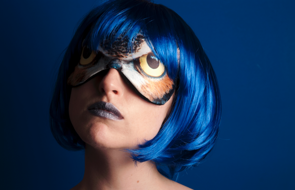 The head of a woman with short and vibrant blue hair is shown wearing a mask over her eyes with the images of owl eyes superimposed over her own eyes