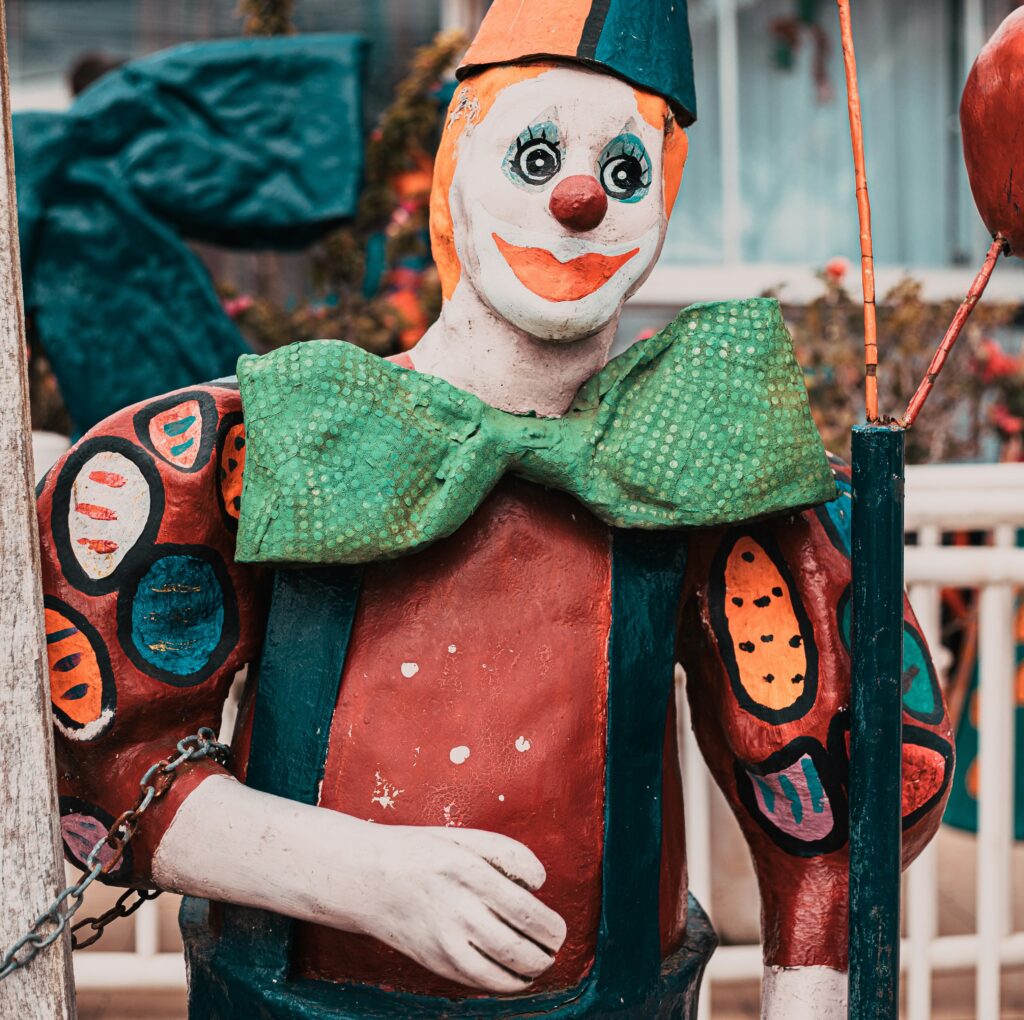 A brightly painted wooden or papier-mache clown with a painted face and huge bowtie stands outside, his right arm chained to a fence