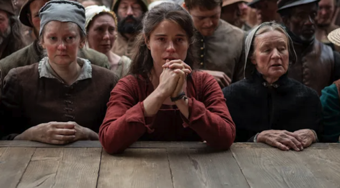 Jessie Buckley as Agnes Shakespeare leans her elbows on a stage between two other women in Elizabethan dress in the film Hamnet