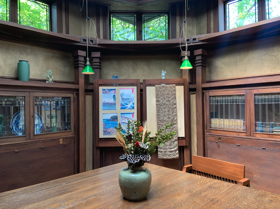A view of part of an octagonal room with windows high up on the walls, small green pendant lights, and glass-fronted cabinets with typical Frank Lloyd Wright-style geometric leaded glass. At front is a green vase on a large oak table that Wright used to display blueprints to his clients. At back is a wall with Japanese prints, figurines, and fabric displayed over it.