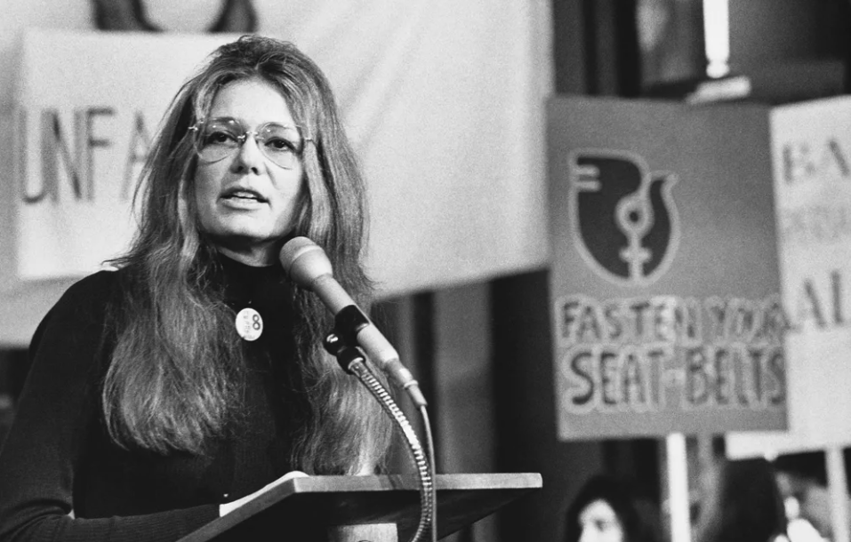 A black and white photo of Gloria Steinem speaking at a microphone at a feminist rally in the early 1970s. She wears her famous aviator wire-rimmed glasses. Behind her are out-of-focus images of women holding up signs on wooden posts. The only sign we can read fully says FASTEN YOUR SEAT-BELTS. The sign features a feminist logo of the Venus sign superimposed on the profile of a dove, which was a well-established symbol of feminism at the time.